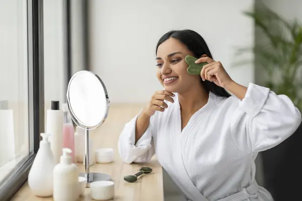 面部按摩的概念 身穿白色浴衣的印度年轻女子坐在浴室的窗边微笑 用绿色的瓜沙 照镜子 享受日常的护肤 — 图库照片