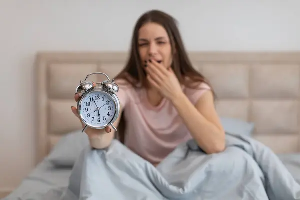 目覚めた若い女性は まだベッドに巣立って 視聴者に向かって古典的な目覚まし時計を拡張し 早起きの普遍的な闘争を伝えます — ストック写真