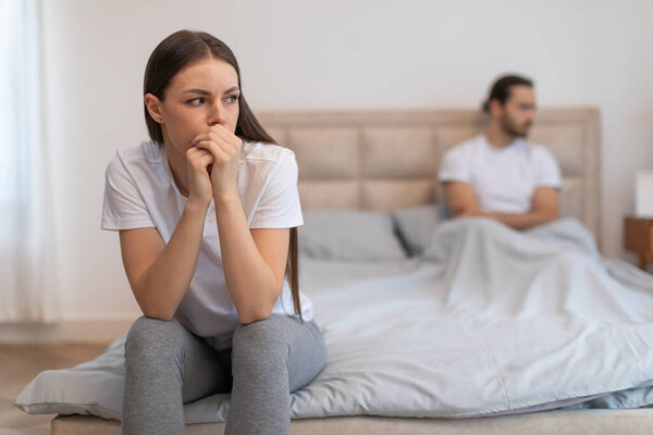 Страстная молодая женщина сидит на переднем плане, руки сжимаются в беспокойстве, в то время как мужчина сидит размытый на заднем плане, изображая напряжение и расстояние в отношениях