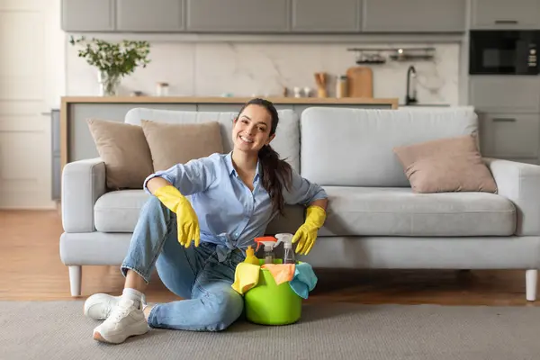 Content woman in casual attire and yellow gloves sitting on the floor, taking break with bucket of cleaning supplies next to her, living room interior, free space