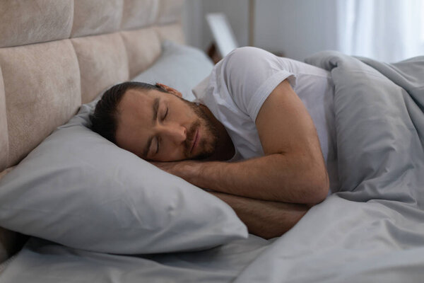 Young man in white t-shirt is in deep sleep, lying on his side on comfortable bedding, illustrating the importance of rest in serene bedroom setting
