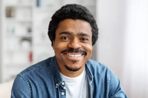 Portrait of cheerful young black man smiling at camera, positive african american male posing indoors in home interior, closeup shot of happy millennial guy in denim shirt, copy space