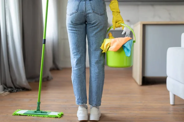 Rear view of woman in casual clothes prepared for cleaning, holding green bucket with cloths and spray bottle, standing beside mop, back view, cropped