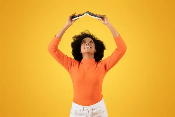 快乐的年轻非洲裔美国女人 卷曲的头发 穿着橙色的高领毛衣和白色的裤子 头顶着一本书 在阳光灿烂的黄色背景下微笑着 工作室 — 图库照片