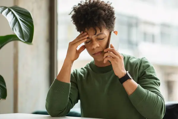 Upset young black guy sitting at table next to window, talking on smartphone, touching his head, have bad news, cafe interior, copy space. Stressed african american businessman have phone conversation