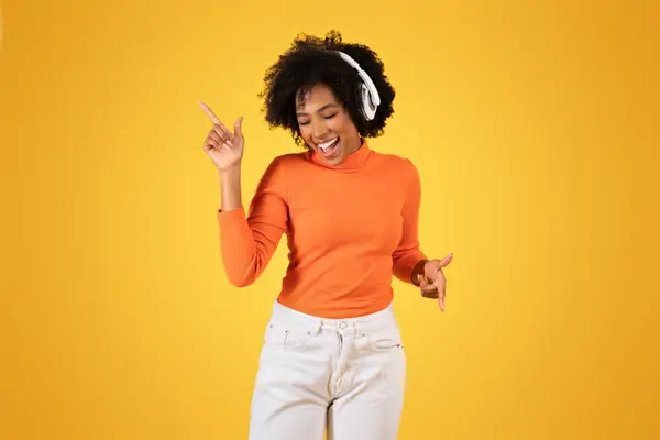 快乐的年轻非洲裔美国女人 卷曲的头发 头戴耳机 头戴橙色毛衣 穿着白色牛仔裤 背景是阳光灿烂的黄色 — 图库照片