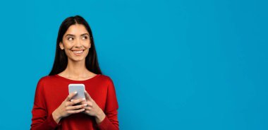 Gülümseyen genç bir kadın akıllı telefonu tutuyor ve fotokopi uzayına bakıyor, mutlu güzel bayan çevrimiçi iletişim için cep telefonunu kullanıyor, mavi stüdyonun arka planında tek başına duruyor, panorama