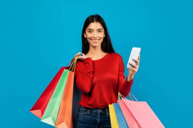 Alışveriş Uygulaması. Güzel genç bayan akıllı telefon ve renkli kağıt alışverişi torbaları, sezonluk indirimler ve teklifler için mobil alışveriş uygulaması kullanan mutlu bayan, mavi arka planda duruyor.