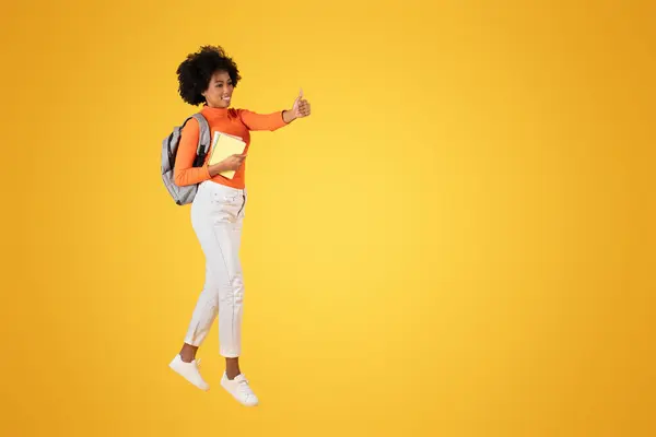 一个乐观的年轻非洲裔美国女人 穿着橙色上衣和白色裤子 拿着笔记本 背着背包 提着大拇指 背景是黄色的 — 图库照片