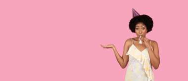 Milenyum boyunca kıvırcık saçlı, parti şapkası takan ve parti kornası çalan, elleri pembe arka planda, stüdyoda şakacı hareketler yapan mutlu Afrikalı Amerikalı kadın.