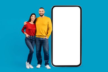 Beyaz ekranlı dev akıllı telefonun yanında duran mutlu genç çift, gülümseyen adam ve kadın mobil uygulama veya web sitesini tanıtıyorlar, ürününüzün reklamını yapıyorlar, mavi stüdyo arka planı,
