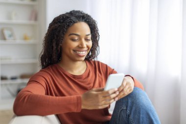 Evdeki koltukta dinlenirken akıllı telefon kullanan gülümseyen siyahi kadın, cep telefonuyla koltukta oturan, yeni uygulamalara ya da internetten alışveriş yapan, oturma odasında dinlenen genç Afrikalı Amerikalı kadın,