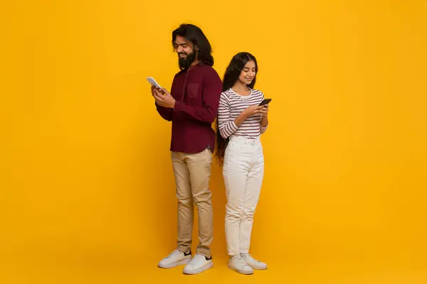 Dating mobile application. Young indian man and woman standing back to back, using smartphone, texting friends, websurfing, scrolling, yellow studio background, copy space, full length