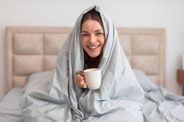 Yumuşak yorgana sarılı neşeli genç bir kadın iki eliyle beyaz kahve fincanı tutuyor. Parlak gülüşü, rahatlamış sabahları sıcaklık ve rahatlık hissi veriyor.