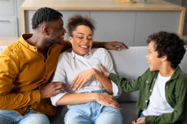 Sevinçli siyah aile, sevgi dolu anın tadını çıkarır, çocuk annesini gıdıklıyor ve gülüyor, neşe dolu etkileşimler kuruyor ve rahat koltukta neşeyi paylaşıyor.