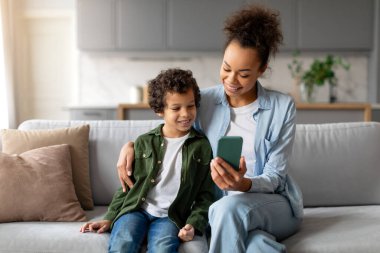 Rahat ve mutlu siyah kıvırcık saçlı genç çocuk ve neşeli annesi birlikte akıllı telefona bakıp rahat bir şekilde kanepede otururken eğlenceli bir anı paylaşıyorlar.