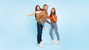 Eğlenceli aile zamanı. Mutlu baba, gülen kızını taşırken annem mavi arka planda birlikte poz vererek barış işareti yapıyor. Ebeveynlerin ve çocuklarının stüdyo fotoğrafları, panorama, kopyalama alanı