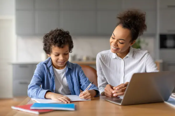 Cheerful black mother with natural curly hair guides her young son as he focuses on writing homework, creating nurturing educational environment at home