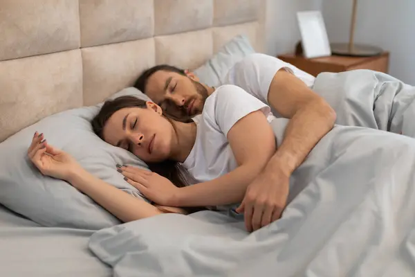 Peaceful couple in white t-shirts, embracing in restful slumber on cozy bed with soft pillows, symbolizing intimacy and comfort in serene bedroom