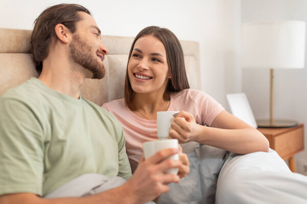 Smiling young couple enjoys cozy moment with coffee cups in bed, sharing warm, relaxed morning together in sunny, intimate bedroom setting and chatting