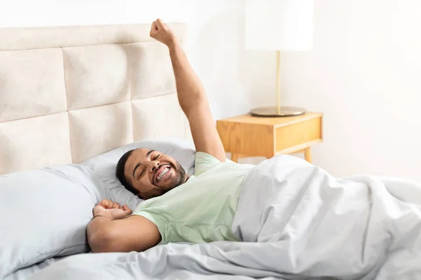 Happy african american bearded guy lying in bed and stretching his body, enjoying good morning, beginning of new day. Young black man slept well, home interior, copy space