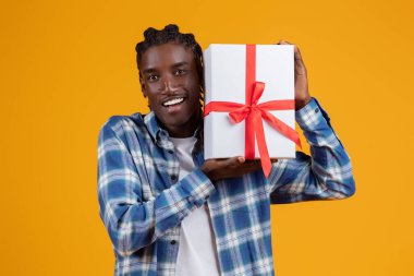 Mutlu genç siyah adam elinde kurdele, neşeli yakışıklı Afrikalı Amerikalı adam hediyesini taşıyor ve şaşırarak ağzını açıyor, sarı stüdyo arka planına karşı duruyor,