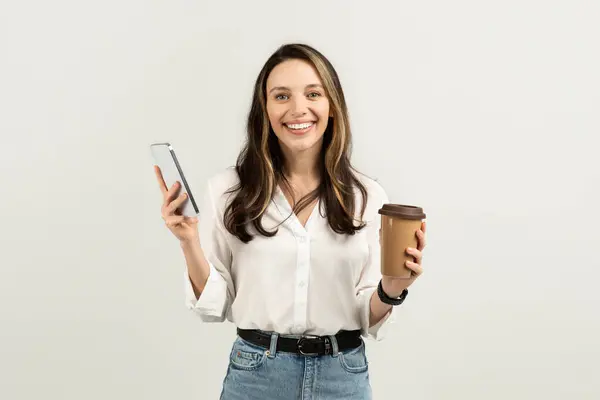 Smiling european woman multitasking with a smartphone in one hand and a coffee cup in the other, dressed in a white blouse and jeans, epitomizing a busy lifestyle, studio