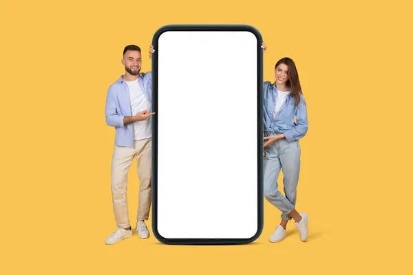 Stock image Smiling man and woman in casual outfits showcasing huge blank smartphone screen, perfect for advertising apps or mobile features, on yellow backdrop, mockup