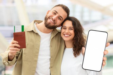 Mutlu Genç Çiftler Seyahat Biletleri ve Havalimanı Terminali 'nde yan yana dururken Akıllı Telefon Gösterimi Neşeli Milenyum Eşleri Yeni Uçuş Rezervasyon Uygulaması ve Mockup Uygulamasını Tavsiye Ediyor