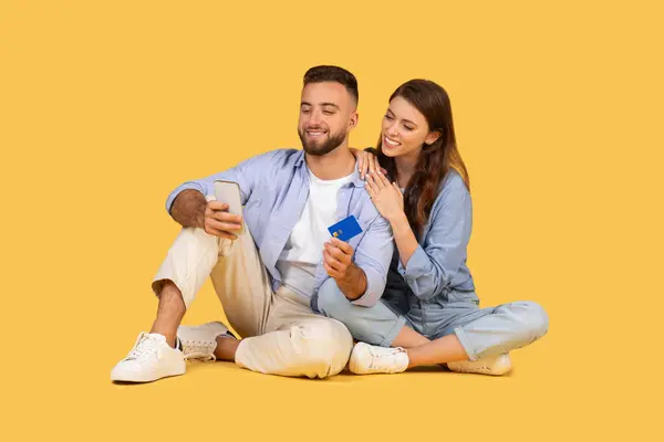 年轻夫妇坐在地板上 男人拿着智能手机和信用卡 男人给女人看屏幕 靠在他身上 一起在黄色背景下购物 — 图库照片