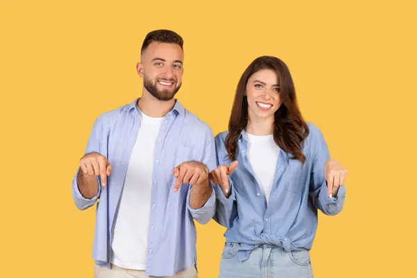カジュアルな青い服を指し下向きを指し 楽しい瞬間を共有し 晴れた黄色の背景に輝く笑顔で楽しい若いユーロペプアのカップル — ストック写真