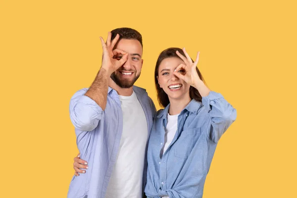 陽気な若いカップルは 眼鏡のような遊び心のある手のジェスチャーを作って 黄色の背景と一緒に光熱した喜びの瞬間を共有します — ストック写真