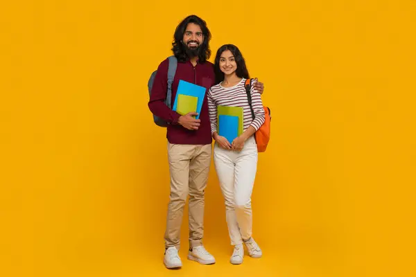 Positive eastern students friends young indian man and woman posing on yellow studio background, holding exercise books and carrying backpacks. College, school, education concept