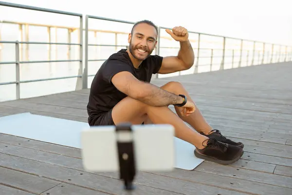 Fit blogger guy in activewear streaming his outdoor workout via smartphone and showing his biceps, bragging about fitness result during training at pier on summer morning