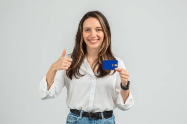Temassız ödeme sembolü olan mavi bir kredi kartı tutan neşeli genç bir kadın, beyaz bluz ve kot pantolon giyiyor, bileğinde akıllı bir bant var.