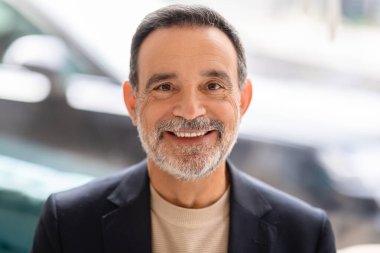 Gri sakallı, parlak gülüşlü, bej bir kazağın üzerine siyah ceket giyen, modern ofis ortamında, aydınlık ve bulanık arka planlı neşeli kıdemli işadamı.