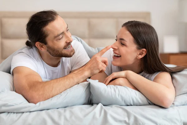 Man playfully touches womans nose as they laugh together, lying in bed, showcasing moment of affection and joyful intimacy in bright bedroom interior