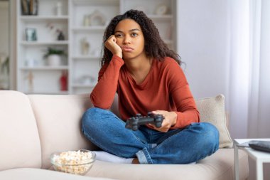 Sıkıcı siyah kadın elinde joystick ile kanepede oturuyor ve başka tarafa bakıyor, evde yalnız başına video oyunları oynuyor, genç Afrikalı Amerikalı kadın kendini yalnız hissediyor, haftasonunu yalnız geçiriyor, boşluğu kopyalıyor.