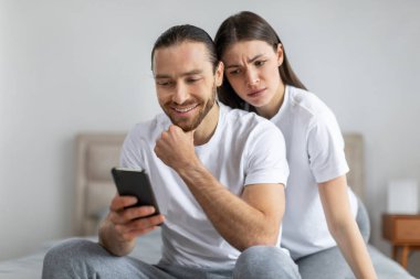 Endişeli bir kadın akıllı telefonuna gülümseyen bir adamın omzundan bakıyor. Yatak odasındaki gizlilik ya da merakı ima ediyor.