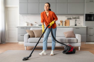 Gündelik giysiler içinde neşeli genç bir bayan ve sarı eldivenler mutlu bir şekilde şık bir oturma odasını süpürüyor parlak kırmızı elektrikli süpürgeyle, gösterişli bir temizlik...