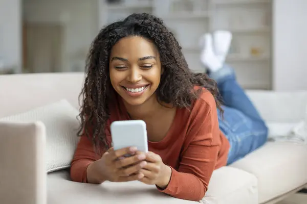 Smiling young black woman texting on smartphone while relaxing at home, happy african american lady using her mobile phone, lying back on couch in cozy living room interior, copy space