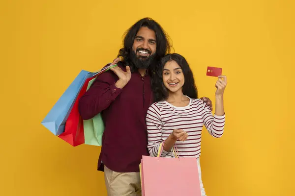 Happy young indian man and woman spouses with colorful paper bags purchases and red plastic bank credit card posing on yellow studio background, recommend easy contactless shopping