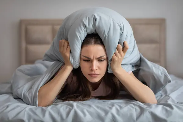 Troubled young woman lying in bed clutches her pillow over her head, her expression one of frustration and annoyance, as she attempts to block out the disruptive noise interrupting her rest