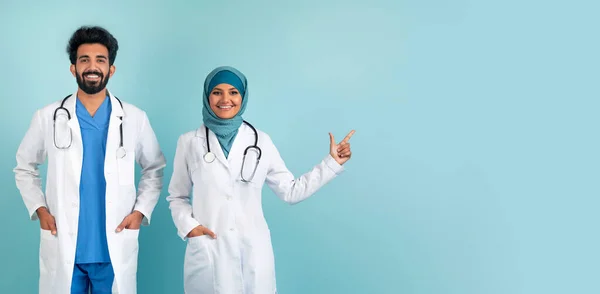 Joyful muslim doctors couple in lab coats pointing aside at copy space, smiling arabic healthcare professionals standing against blue studio background, exemplifying teamwork and positivity, panorama