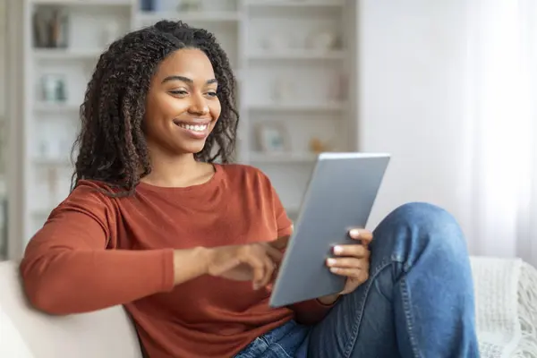 Portrait Of Young Smiling Black Woman Resting On Couch With Digital Tablet, Happy African American Female Browsing Internet Or Shopping Online While Relaxing With Tab Computer At Home, Copy Space
