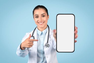 Neşeli bayan doktor boş akıllı telefon ekranına işaret ediyor, tıbbi uygulama veya web sitesi görüntüsü için ideal, üniformalı ve steteskoplu bayan açık mavi arka plan,