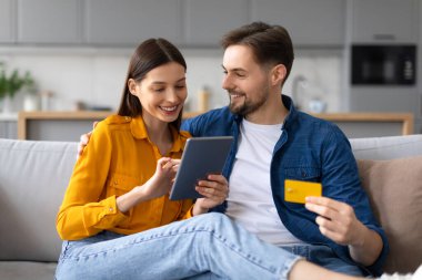 Neşeli, genç eşler, kredi kartı olan bir adam ve tablet kullanan bir kadın, online alışveriş yapıyor, modern oturma odasındaki rahat kanepeye yakın oturuyor.