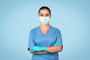 Mavi önlüklü profesyonel bayan hemşire, cerrahi maske ve kolları çapraz duran eldivenler, sakin mavi arka planda tıbbi görevlere hazır.