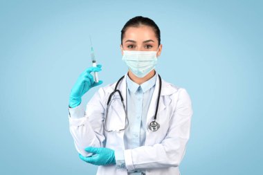 Şırınga tutan, maske takan, steril eldiven takan, tıbbi prosedüre hazır, soluk mavi arka planda poz veren kadın doktor.