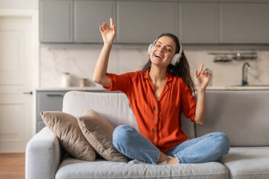 Canlı turuncu gömlekli neşeli genç bir kadın kulaklıklarıyla müzikle kaybolmuş, evinde rahat bir koltukta otururken neşeyle dans ediyor.